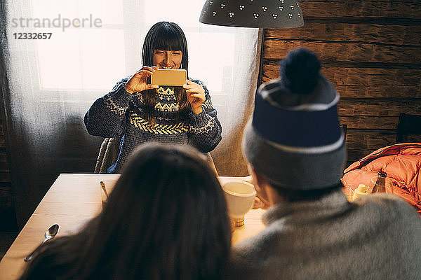 Woman photographing friends through mobile phone while sitting in log cabin