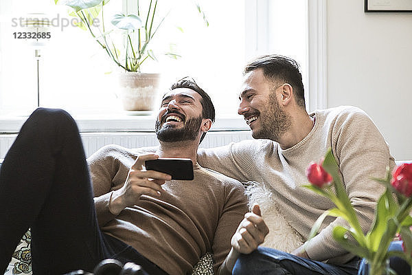 Cheerful gay couple with smart phone sitting on sofa against window at home