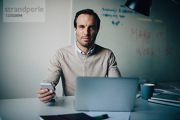 Portrait of confident businessman sitting with smart phone and laptop at desk in creative office