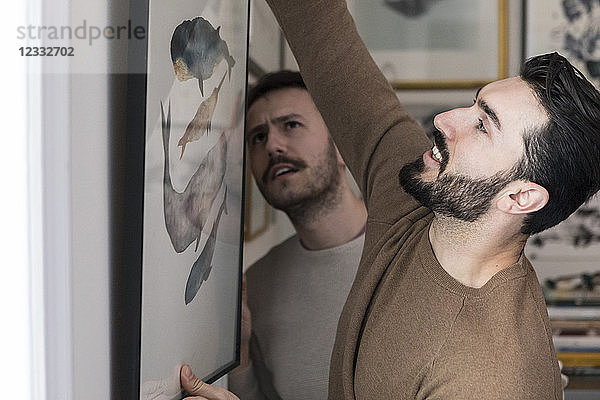 Young gay couple hanging painting on wall at home