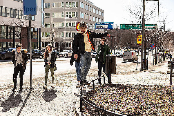 Teenage boy balancing on railing while friends walking on sidewalk in city during sunny day