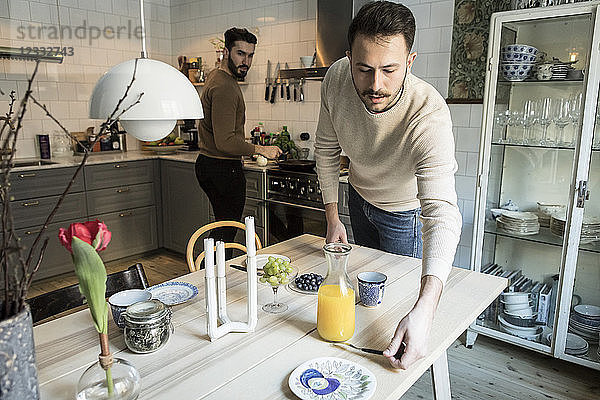 Young man setting table while partner working in background at kitchen