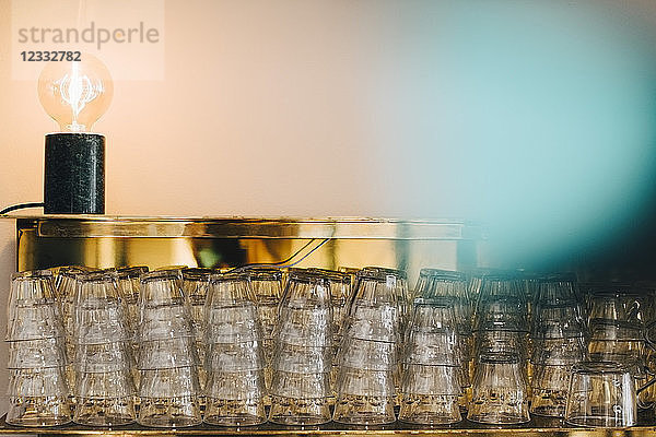 Stack of drinking glasses on shelf by illuminated light bulb against wall in office canteen