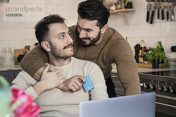 Romantic gay couple doing online shopping through laptop in kitchen at home