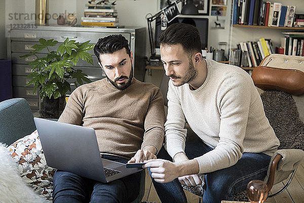 Gay couple shopping online with credit card through laptop while relaxing on chairs at home
