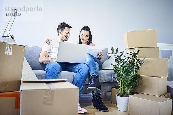 Happy couple sitting on couch surrounded by cardboard boxes using laptop