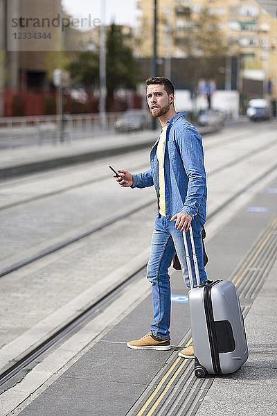 Young man waiting at a station with smartphone in his hand and trolley