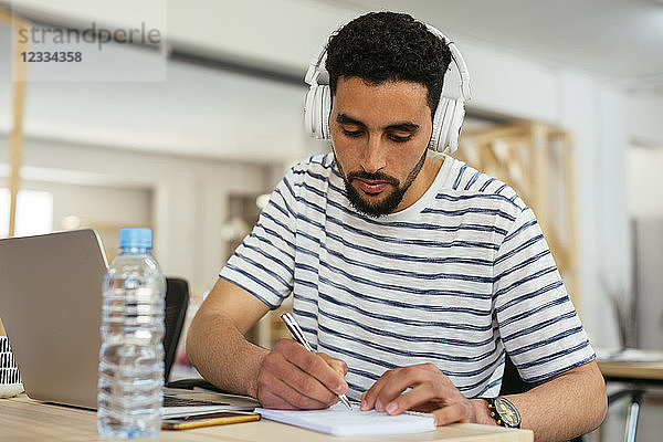 Young man wearing headphones taking notes at desk in office