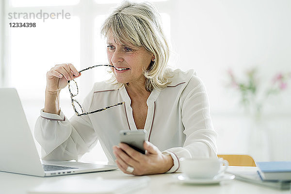 Mature businesswoman holding cell phone working on laptop at desk