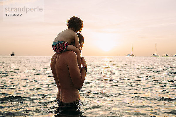 Back view of father carrying his little daughter on shoulders in the sea watching sunset