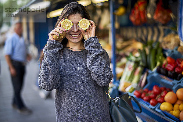 Laughing young woman on market covering her eyes with lemon halves