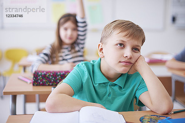 Bored schoolboy in class with girl raising her hand in background