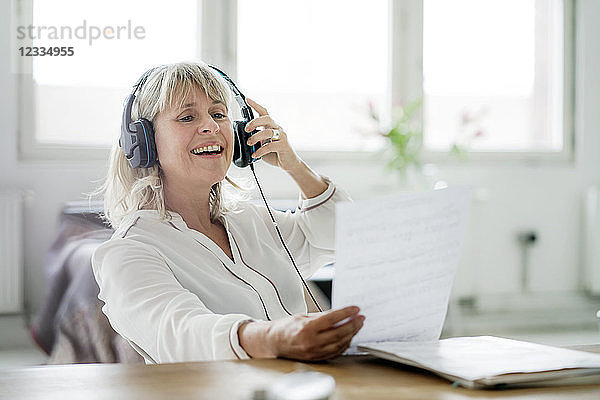 Laughing mature businesswoman wearing headphones looking at document at desk