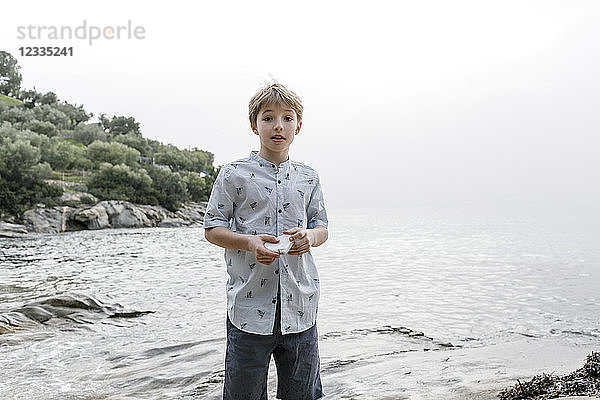 Greece  Chalkidiki  portrait of blond boy standing in front of the sea holding stone