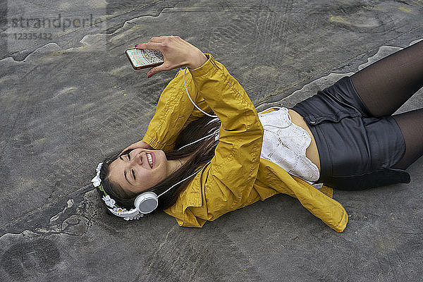 Smiling woman with headphones lying on ground taking selfie with smartphone