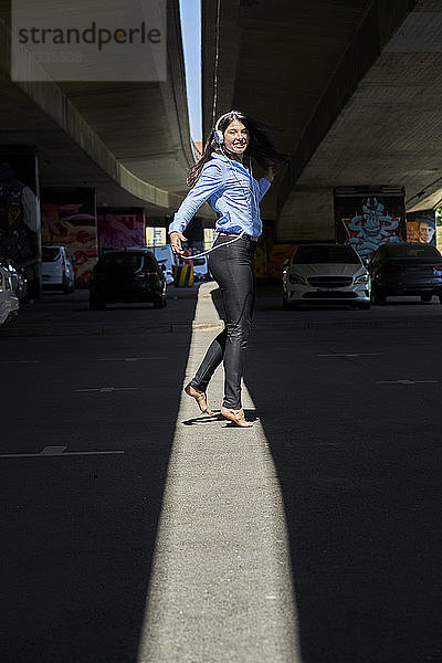 Cheerful young woman with headphones and cell phone dancing barefoot at underpass