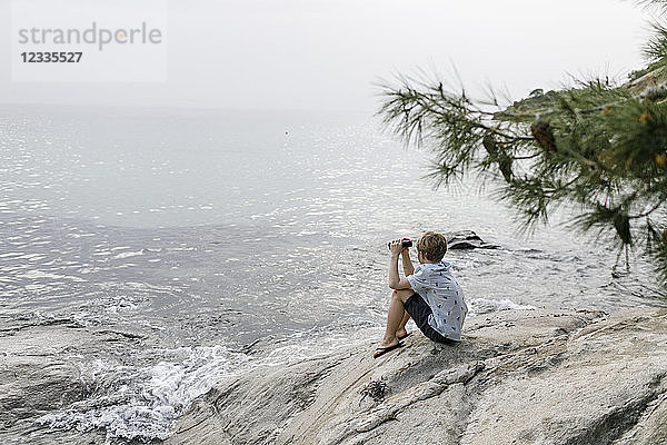 Greece  Chalkidiki  boy with binoculars sitting on rock looking at the sea