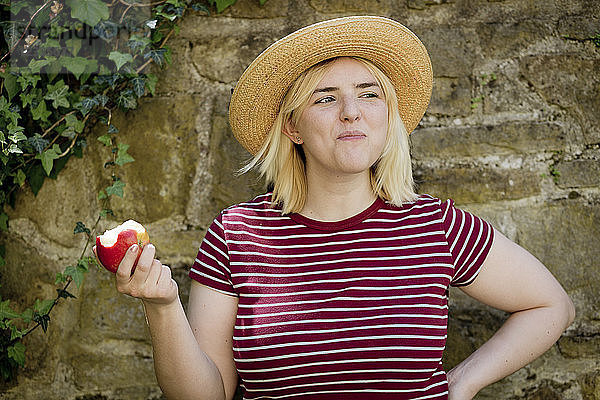 Portrait of young blonde woman with sun hat eating apple