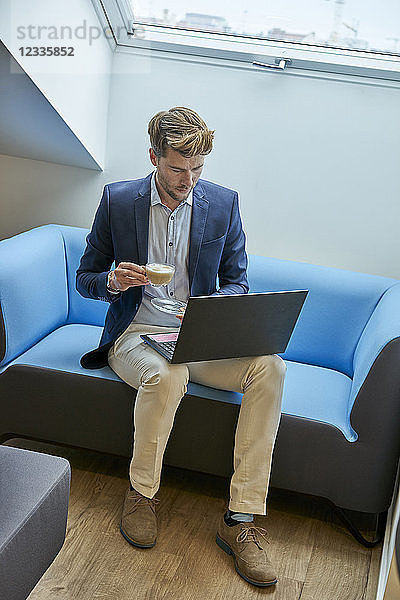 Businessman sitting on couch with cup of coffee looking at laptop