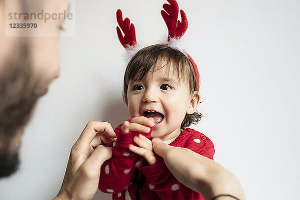 Portrait of laughing baby girl with reindeer antlers headband getting dressed by her father
