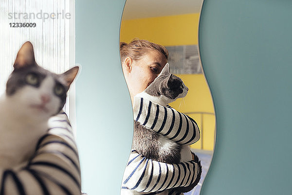 Woman holding a gray cat next to a mirror at home