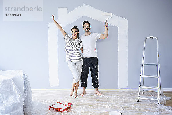 Portrait of cheerful couple painting in new apartment with house shape on wall