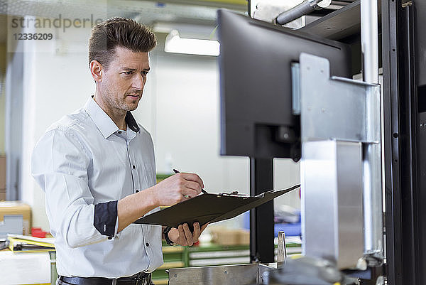 Man with clipboard in factory looking at screen