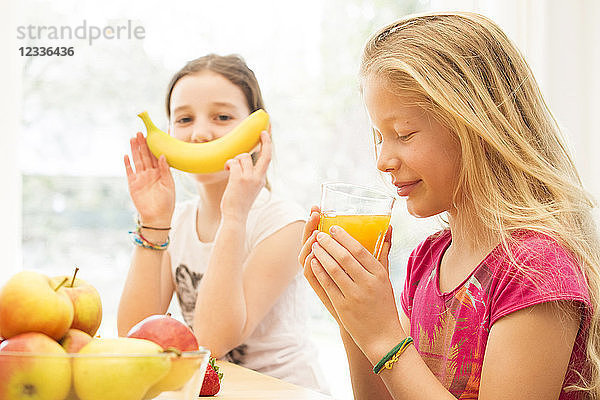 Portrait of girl drinking glass of orange juice while her friend having fun with banana