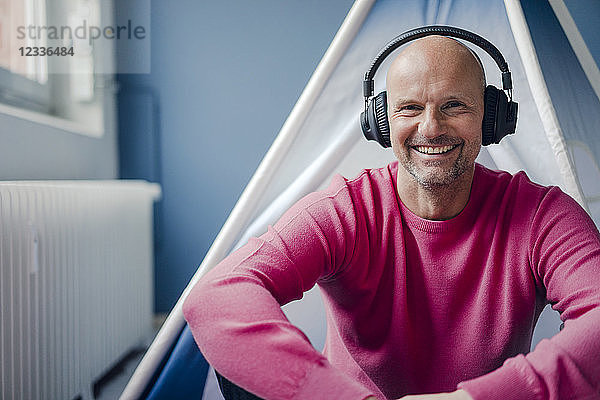 Portrait of smiling mature man wearing headphones sitting at teepee indoors