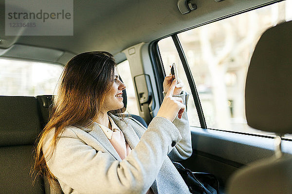 Smiling young woman sitting on backseat of a car taking picture with cell phone