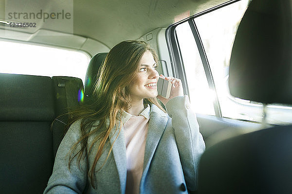 Smiling young businesswoman on the phone sitting on backseat of a car looking out of window