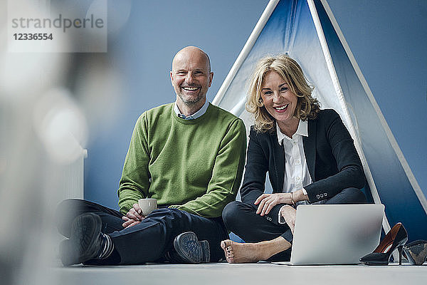 Happy businessman and businesswoman sitting at teepee indoors with laptop