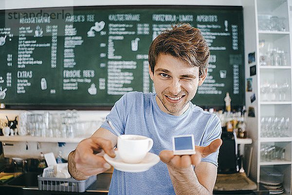 Smiling man in a cafe offering cup of coffee and holding miniature laptop model