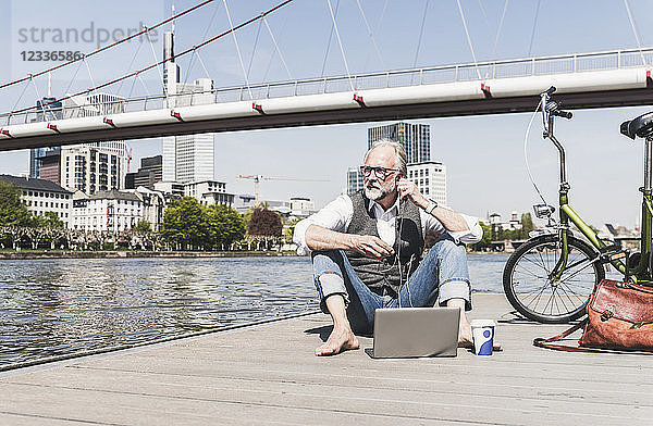 Mature man with laptop  earbuds and bicycle sitting at the riverside in the city