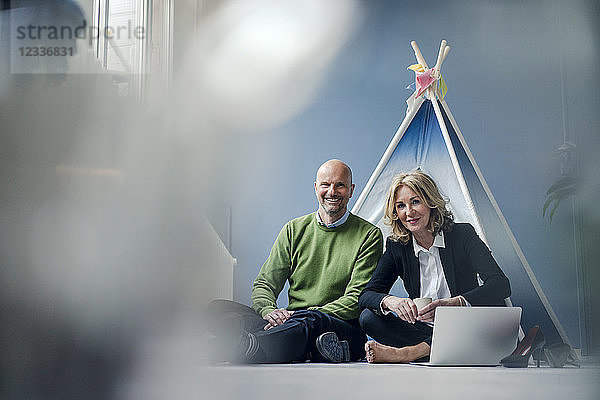 Smiling businessman and businesswoman sitting at teepee indoors with laptop