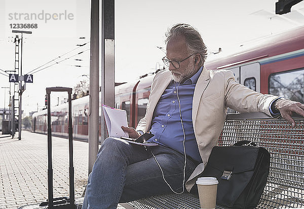Mature businessman sitting at train station with cell phone  earbuds and notebook
