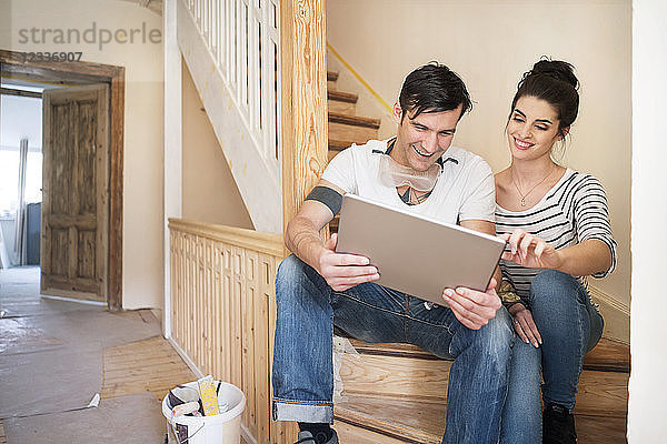 Young couple refurbishing new home  sitting on stairs using digital tablet