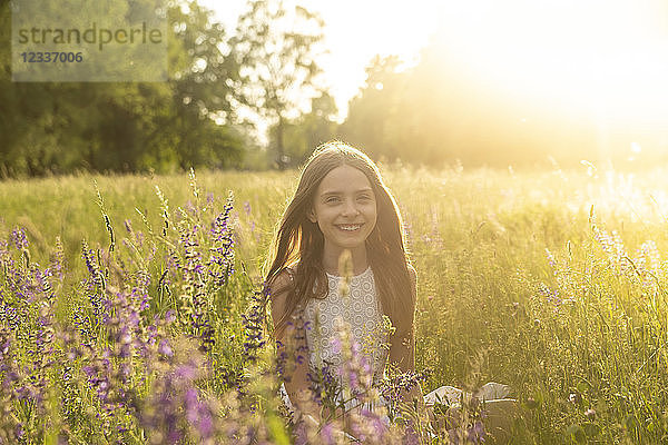 Portrait of smiling girl crouching on flower meadow at evening twilight