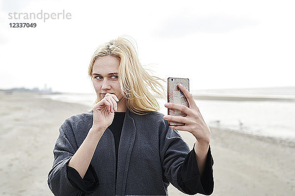 Netherlands  portrait of blond young woman taking selfie with smartphone on the beach