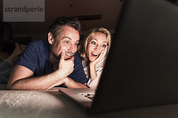 Amazed mature couple lying on couch at home looking at laptop