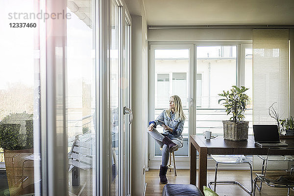 Woman with tablet sitting on stool looking out of window