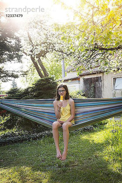Smiling girl sitting on hammock in the garden eating ice lolly