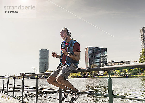 Happy athletic mature man with headphones sitting at the riverside in the city