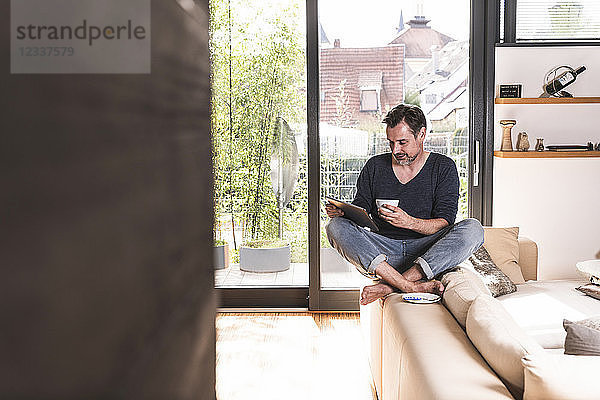 Mature man with cup of coffee sitting on backrest of couch using tablet