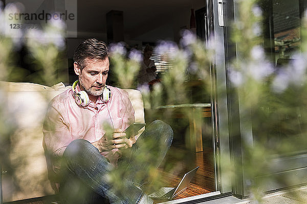 Mature man sitting at open terrace door using smartphone