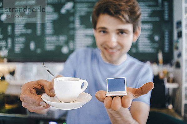Smiling man in a cafe offering cup of coffee and holding miniature laptop model