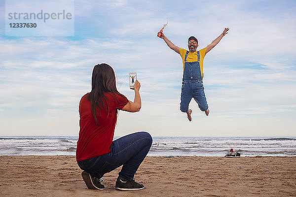 Woman taking smartphone picture of happy man jumping on the beach