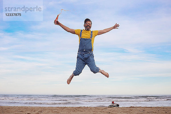 Portrait of happy man in dungarees jumping with soda in hand on the beach
