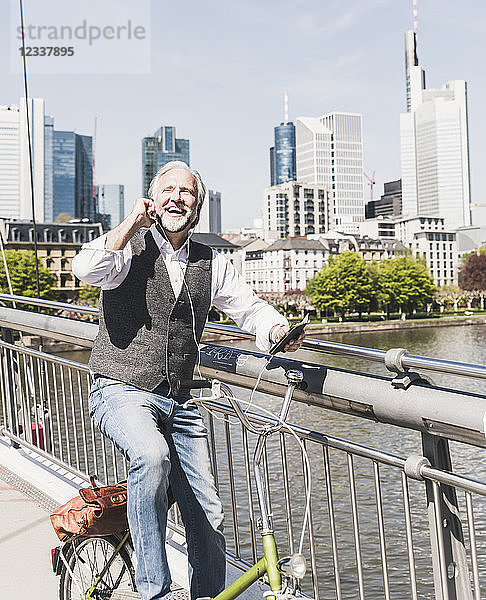 Happy mature man with earbuds  cell phone and bicycle on bridge in the city