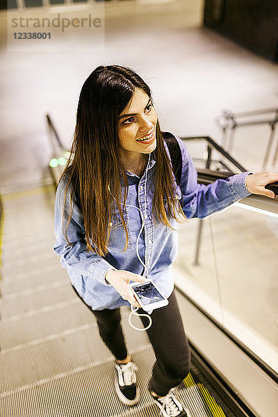 Portrait of smiling young woman standing on escalator listening music with cell phone and earphones